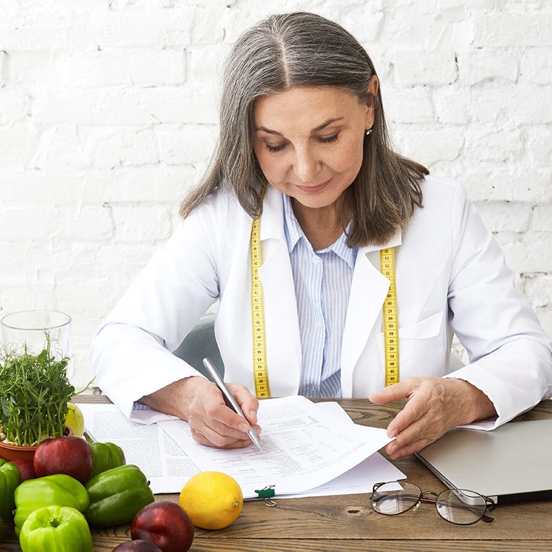 Hardworking experienced Caucasian elderly female nutritionist wearing white gown and measuring tape around her neck, writing medical records and prescriptions, sitting at desk with fresh vegetables